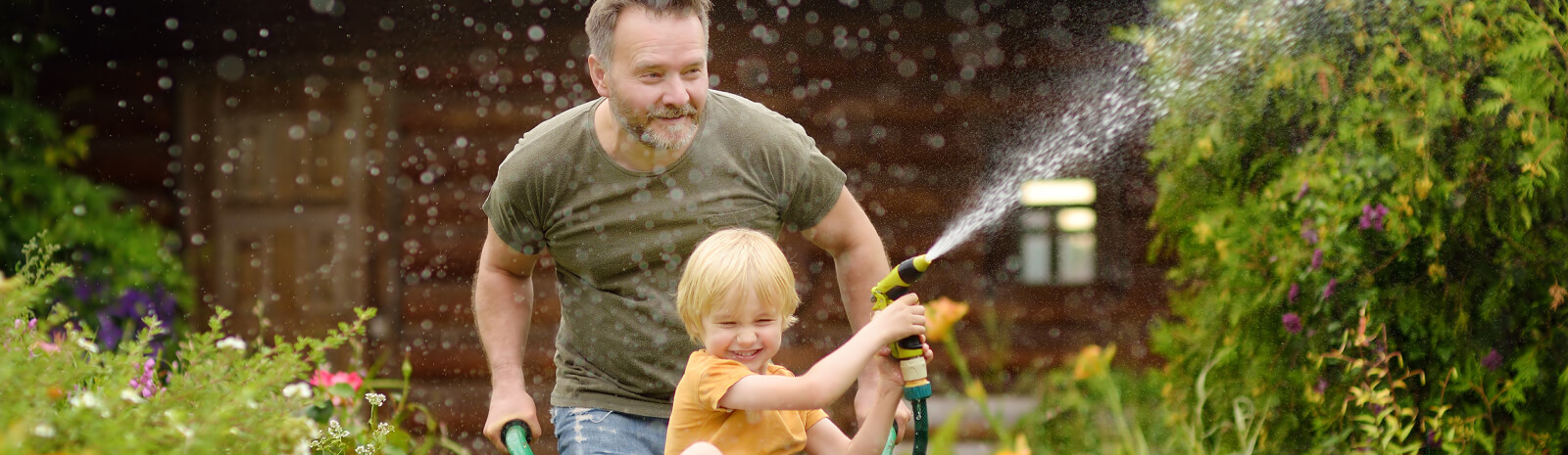 Dad pushing young kid in a wheelbarrow while holding a water hose