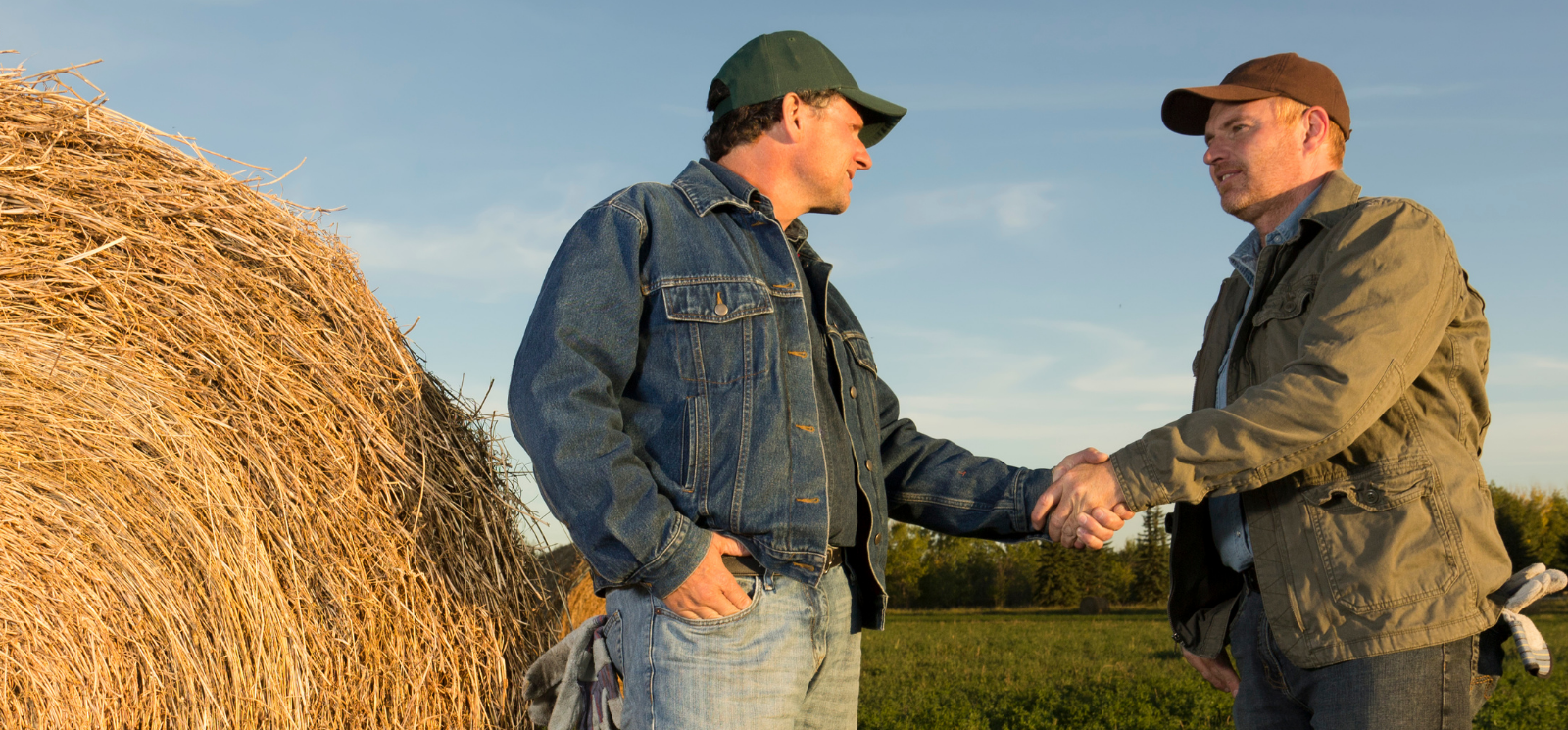 two men wearing baseball caps, jean jacket, and work jacket, and jeans, shaking hands in front of a round bale and a field.